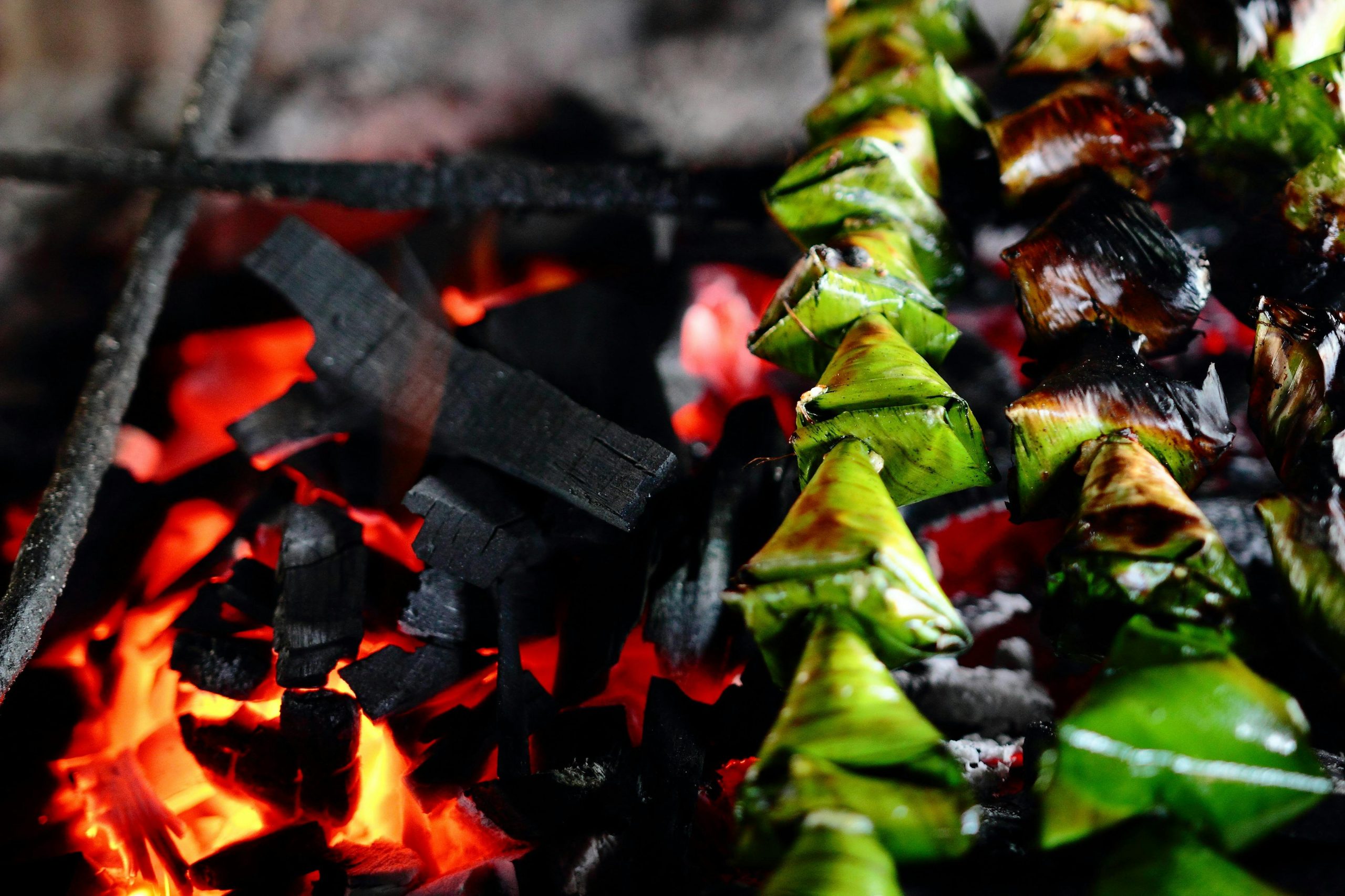 Close-up of traditional Malaysian snacks wrapped in leaves being grilled over glowing charcoal embers.